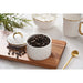 A white small canister with a diamond-quilted pattern and gold trim, shown open with coffee beans inside on a wooden board. A matching lid sits beside it, with a gold spoon and other white-and-gold tableware in the background.