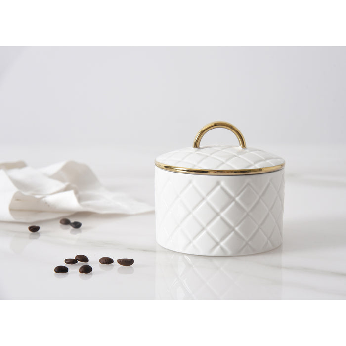 A white small lidded container with a quilted diamond pattern and a gold-tone handle, set on a white marble surface with scattered coffee beans and a crumpled napkin in the background.