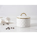 A white small lidded container with a quilted diamond pattern and a gold-tone handle, set on a white marble surface with scattered coffee beans and a crumpled napkin in the background.