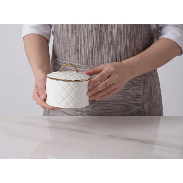 A person wearing a gray striped apron holds a white small lidded container with a quilted pattern and gold trim on a light marble countertop.