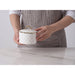 A person wearing a gray striped apron holds a white small lidded container with a quilted pattern and gold trim on a light marble countertop.