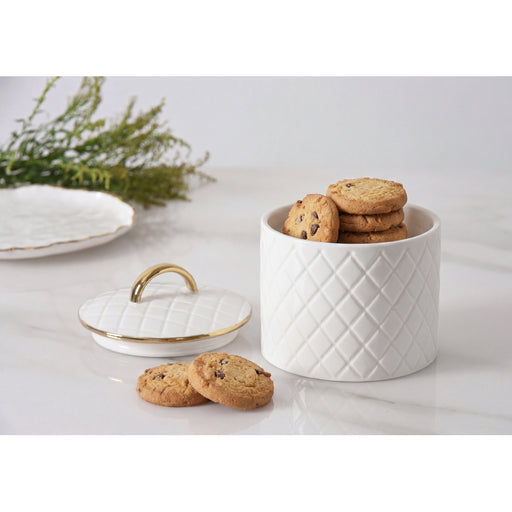 A white cookie jar with a diamond-quilted pattern and a gold-trimmed lid, shown on a light marble surface with cookies inside and a few placed in front. A blurred plate and greenery appear in the background.