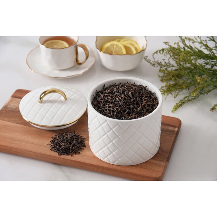 A white tea canister with a diamond-quilted pattern and gold trim, shown open with loose-leaf tea inside on a wooden board, alongside a matching lid and teacups in the background.