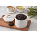 A white tea canister with a diamond-quilted pattern and gold trim, shown open with loose-leaf tea inside on a wooden board, alongside a matching lid and teacups in the background.