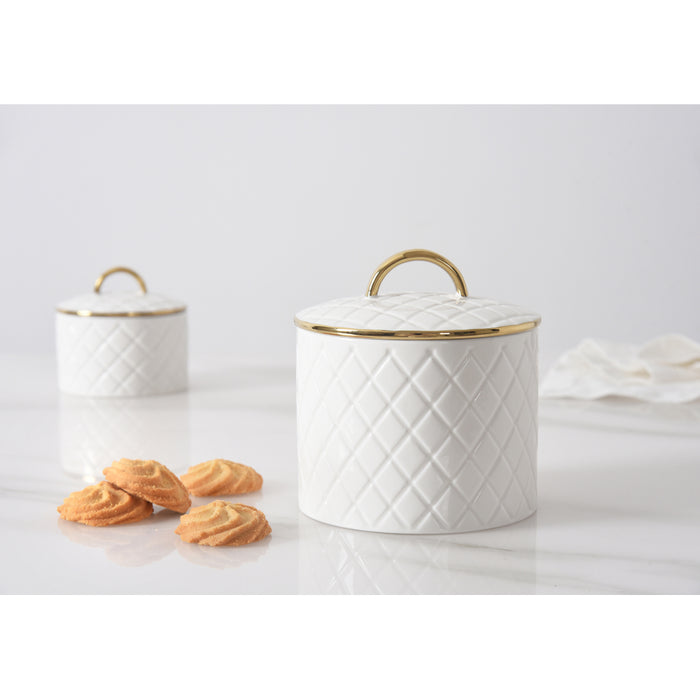 Two white lidded canisters with a quilted diamond pattern and gold trim, shown on a light marble surface with a few cookies in front and a white cloth on the right.