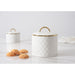 Two white lidded canisters with a quilted diamond pattern and gold trim, shown on a light marble surface with a few cookies in front and a white cloth on the right.