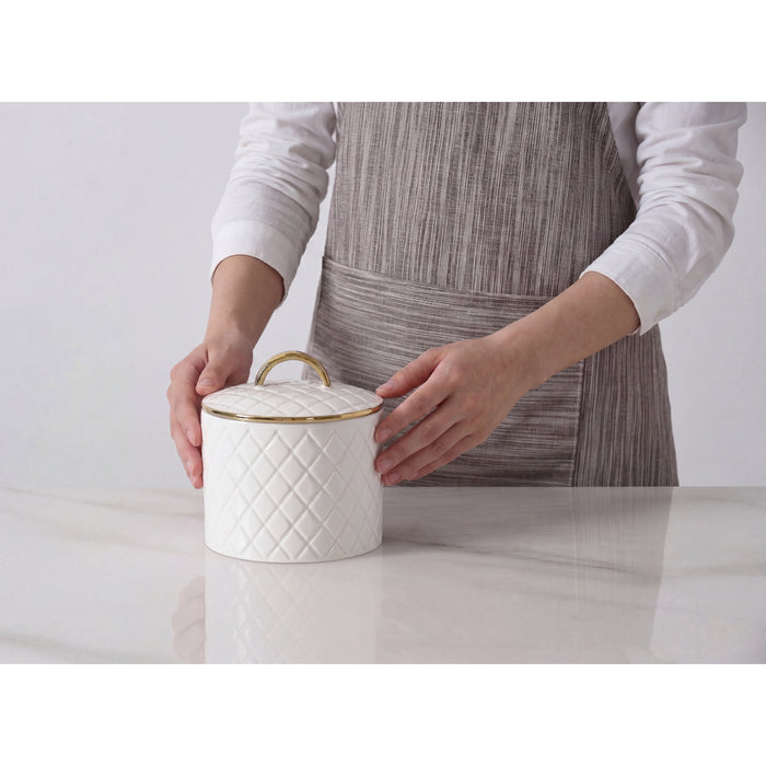 A white lidded container with a quilted diamond pattern and gold trim, held by a person wearing a gray striped apron over a white long-sleeve shirt on a light gray background.