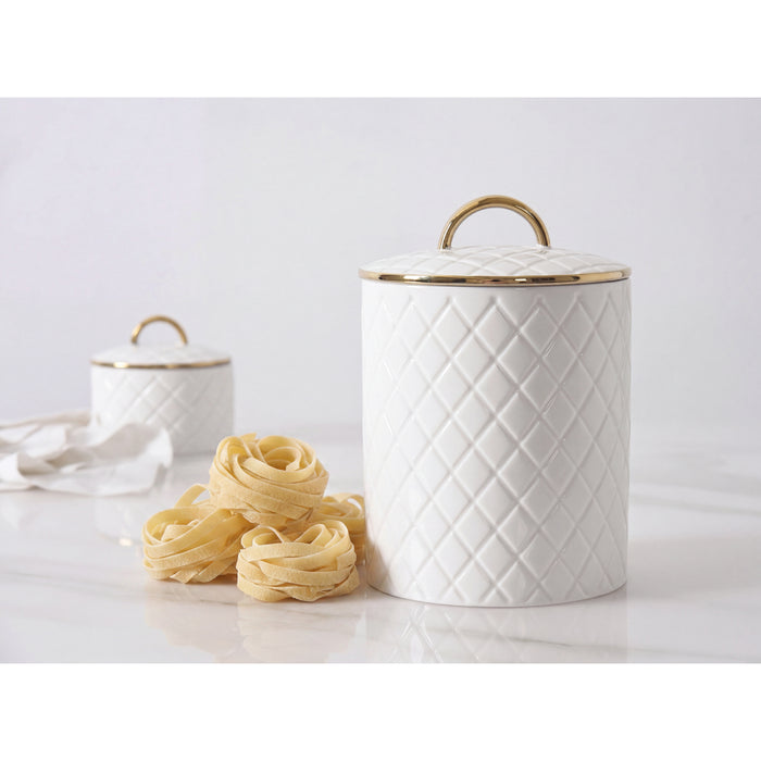 A white large canister with a diamond-quilted pattern and gold trim, shown with a matching lid and handle. A small stack of dried pasta sits in front on a light marble-like surface, with a second matching canister blurred in the background.