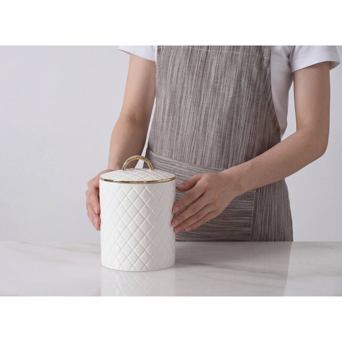 A person wearing a gray striped apron holds a white quilted large canister with a lid and gold trim on a light marble surface against a white background.