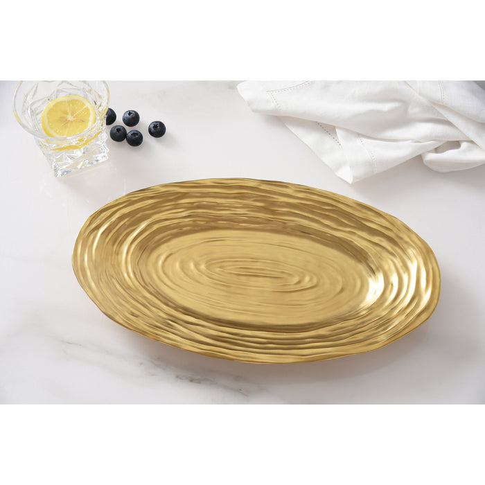 An oval gold serving platter with a textured, concentric-ring pattern, styled on a white marble surface with a glass of lemon water, blueberries, and a white napkin.
