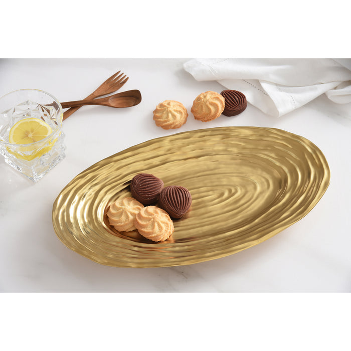 An oval gold serving platter with a textured concentric-ring pattern, styled on a white marble surface with cookies, a glass of lemonade, and wooden cutlery.