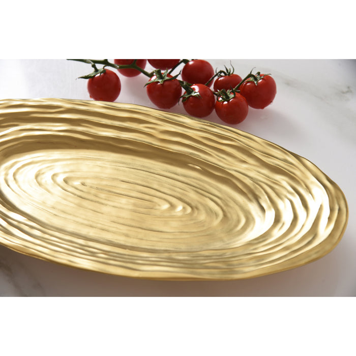 A gold oval serving platter with concentric textured rings, shown on a white marble surface with a small cluster of red cherry tomatoes in the background.