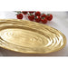 A gold oval serving platter with concentric textured rings, shown on a white marble surface with a small cluster of red cherry tomatoes in the background.