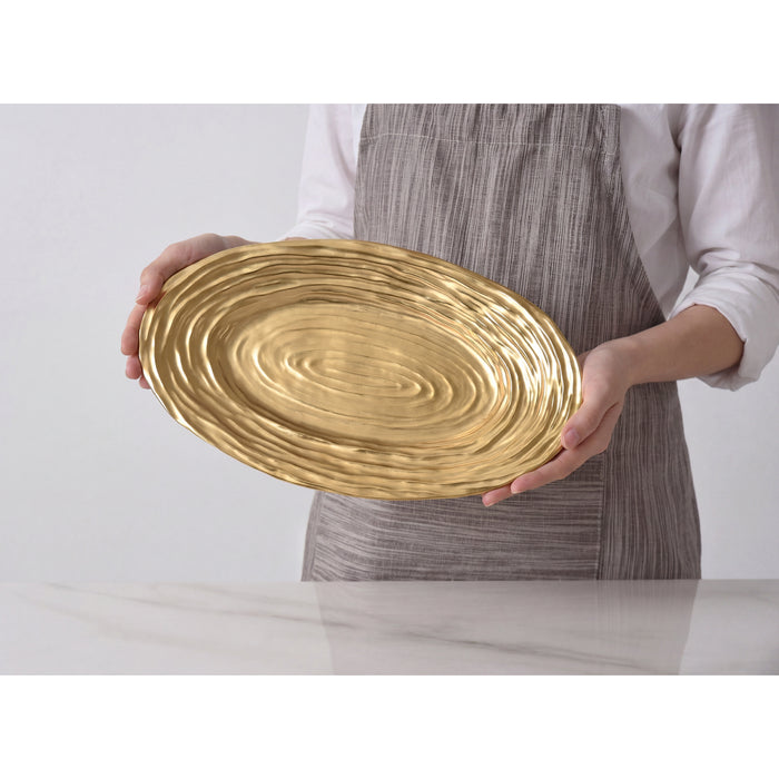 A person wearing a white shirt and gray striped apron holds an oval gold platter with a textured, concentric-ring finish over a white marble surface.