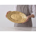 A person wearing a white shirt and gray striped apron holds an oval gold platter with a textured, concentric-ring finish over a white marble surface.