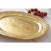 A large oval gold serving platter with a textured, concentric-ring pattern, shown on a white marble surface next to a small bunch of red cherry tomatoes.