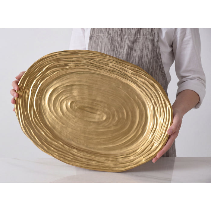 A person in a white shirt and gray striped apron holds a large oval gold serving platter with a textured pattern against a clean white background.