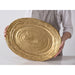 A person in a white shirt and gray striped apron holds a large oval gold serving platter with a textured pattern against a clean white background.