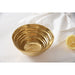 A gold bowl with a concentric wave texture, styled on a white surface next to a white napkin and a lemon slice.