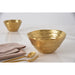 A gold bowl is shown prominently on a white surface, with matching gold cutlery and a folded blush pink napkin in the foreground. A second matching bowl appears softly blurred in the background.