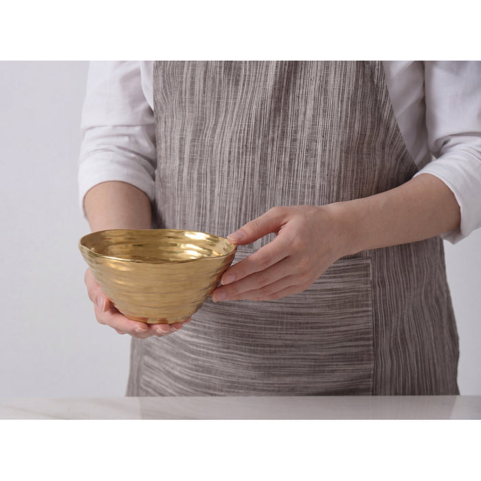 A person wearing a white shirt and a gray striped apron holds a small, textured gold bowl against a light gray background.