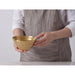 A person wearing a white shirt and a gray striped apron holds a small, textured gold bowl against a light gray background.