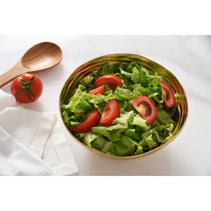 A gold serving bowl filled with a fresh green salad of lettuce and tomato slices, styled on a white marble surface with a wooden spoon, a whole tomato, and a white napkin.