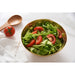 A gold serving bowl filled with a fresh green salad of lettuce and tomato slices, styled on a white marble surface with a wooden spoon, a whole tomato, and a white napkin.