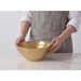 A person wearing a white shirt and gray apron holds a large gold bowl on a white marble-like surface against a light gray background.