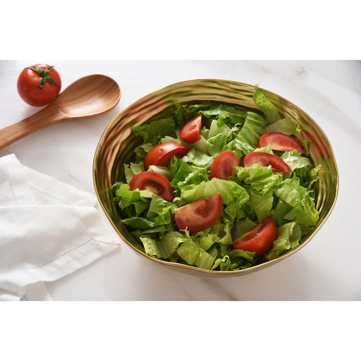 A large gold salad serving wavy bowl filled with a green salad of lettuce and tomato slices, styled on a white marble surface with a wooden spoon, a cherry tomato, and a white napkin.