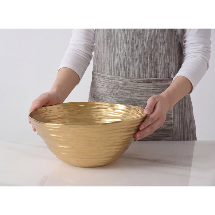 A person wearing a gray striped apron and white long-sleeve shirt holds a large gold salad serving bowl on a white marble countertop against a light gray background.