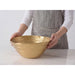 A person wearing a gray striped apron and white long-sleeve shirt holds a large gold salad serving bowl on a white marble countertop against a light gray background.